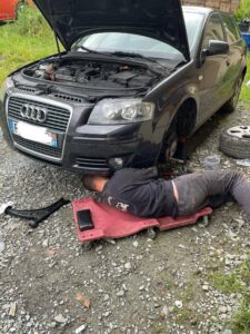 réparation d’une Audi chez amlauto à Cadalen, mécanicien sous le véhicule, outils et roue déposée sur graviers,